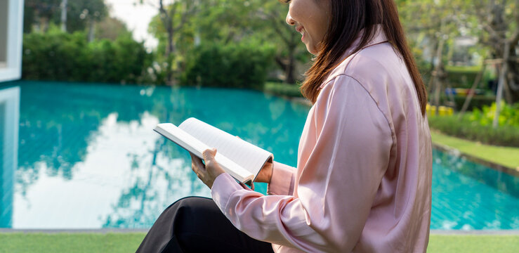 Woman reading notebook by pool in garden. Relaxed woman wearing silk blouse and black pants enjoying quiet outdoor reading and note-taking by a blue pool in a lush garden on a sunny afternoon.