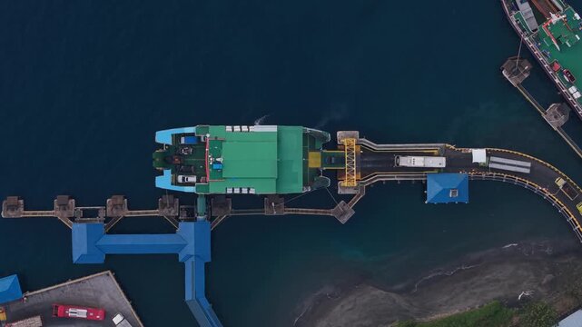 Aerial view of Gilimanuk Port, calm blue waters. Green container ship docked near blue loading platforms. Large yellow crane stands ready for cargo operations. Workers move quietly