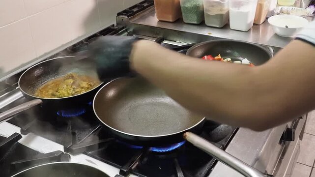Indian chef sauteing mixed vegetables in pan beside another cooking preparation on commercial gas range, showing professional restaurant kitchen food preparation