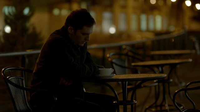 Young man sits at an outdoor cafe table beside a white cup, glancing across the warmly lit terrace before lowering his gaze as empty chairs and soft city lights blur behind him