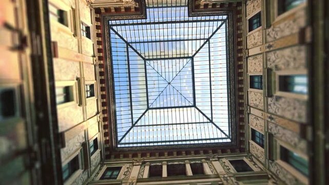 Direct upward view of a geometric glass and iron skylight revealing blue sky above ornate historic balconies and decorative walls inside Galleria Subalpina in Turin Italy.