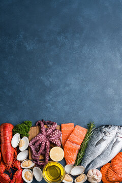 A flat lay image of raw seafood arranged on a grey slate background. Red lobster, green parsley, clam shells, octopus, olive oil and salmon.