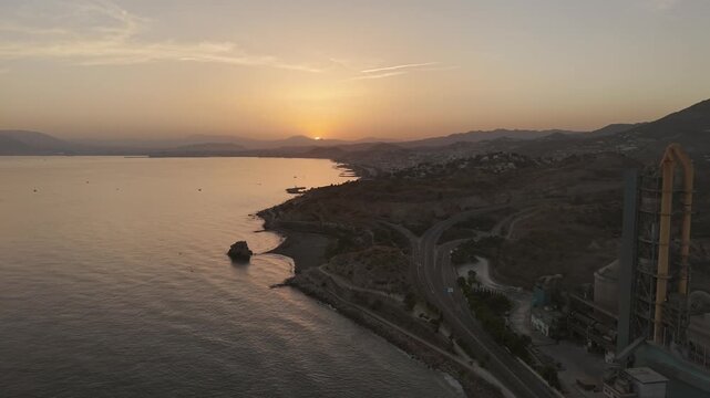 Elevated drone shot at Playa Penon del Cuervo shoreline with regional highway and La Arana cement plant stretch along the calm sea during sunset, Malaga, Andalusia, Spain