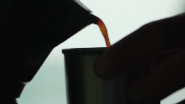 Dark espresso pouring from a metal moka pot into a small vessel inside a brightly lit room. Static close-up shot.