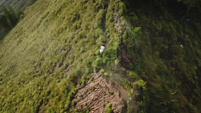 Man running narrow path on steep, grassy mountain ridge, mist clinging over green valley below, representing adventure, exploration, nature, travel. Aerial view tracking flight. Batur, Bali, Indonesia