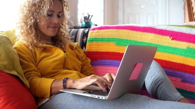A happy curly-haired woman relaxes on a vibrant sofa at home, casually browsing on her laptop with a smile, embracing modern leisure and digital connection in a comfortable setting.