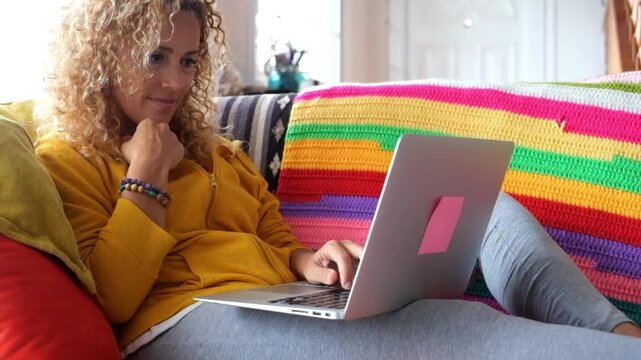 A happy curly-haired woman relaxes on a vibrant sofa at home, casually browsing on her laptop with a smile, embracing modern leisure and digital connection in a comfortable setting.