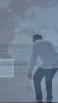 Vertical video: Senior man walking to shore crouching, touching sand causing tech UI overlaying map