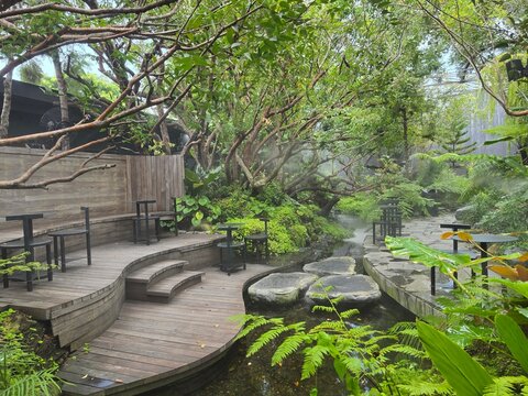 A lush tropical garden cafe with curved wooden decks, black tables, and a misty stream crossed by stepping stones amid dense greenery.