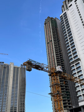 A construction site featuring two tall buildings in progress. A yellow crane is prominently positioned, working on the left structure. The sky is clear and blue.