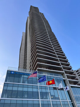 A tall modern skyscraper stands against a clear blue sky. The building features a glass facade and numerous balconies. Flags of various countries are displayed in front.