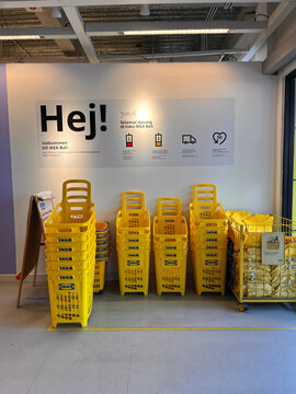 A bright store interior features a large wall with the word 'Hej!' displayed prominently. Yellow shopping baskets are stacked neatly on the floor. The area exudes a welcoming atmosphere.