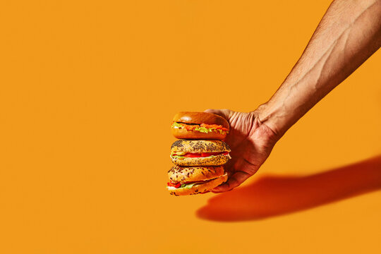 Hand Holding a Stack of Fresh Bagel Sandwiches on Orange Background