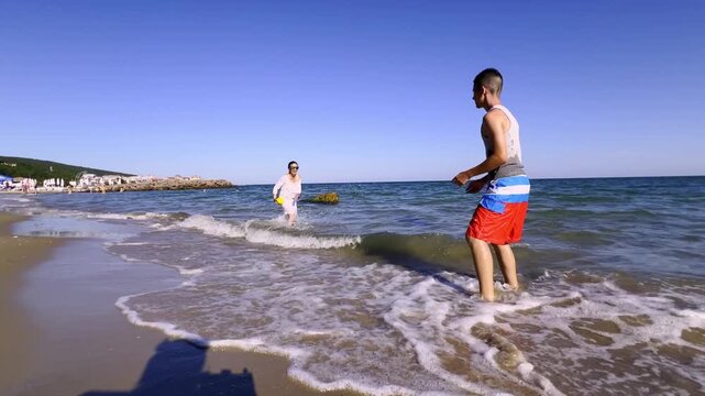 Mature mom and teenage son splashing water, laughing, and fooling around with ball on sunny ocean beach getaway.. High quality FullHD footage