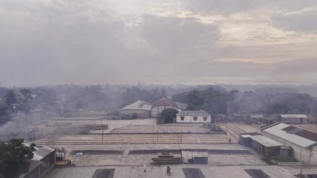 Backward drone footage pulls away from a Khajanagar rice mill drying yard, revealing workers manually handling paddy as early morning rice processing begins at dawn in rural Bangladesh.
