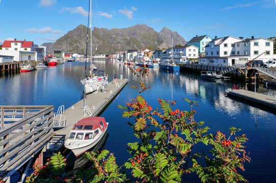 Sunny harbor scene in Henningsv&aelig;r on the Lofoten Islands with sailboats, motorboats, colorful houses and calm water against an impressive rocky mountain landscape under blue sky.