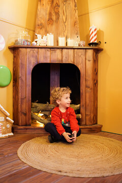 Happy curly haired boy sitting on rug in front of wooden fireplace decorated for Christmas