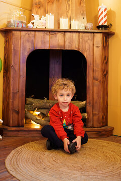 Little boy wearing a Christmas sweater sitting on rug by fireplace, celebrating winter holidays