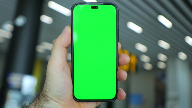 Hand holds phone with green screen. Background shows airport terminal. Terminal has overhead lights and gate signs. Green screen is plain, no content. Focus on phone, background slightly blurred