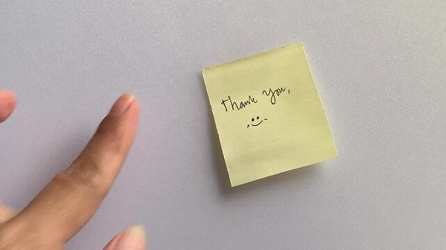 Two hands sticking a yellow note with a handwritten says thank you  onto a white wall. Close-up static view in bright daylight.