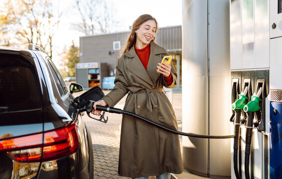Young woman stands at gas station with phone in hand, refueling her car. Woman with a smartphone at a gas station at sunset. Concept of transportation, refueling, and technology.