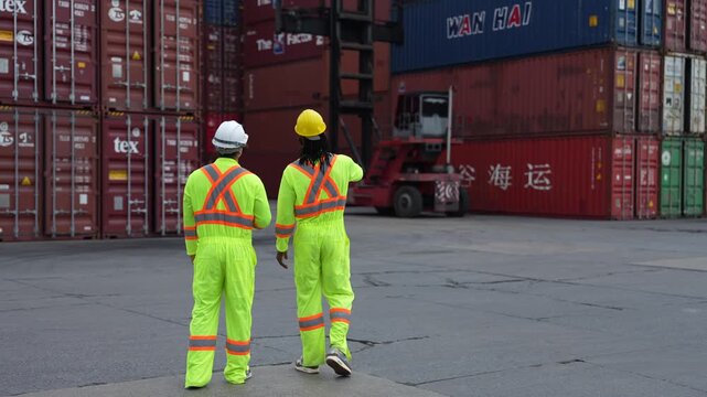 Two dock workers in high visibility coveralls walk away from camera across the container yard toward a working reach stacker during a routine shift at the container terminal.
