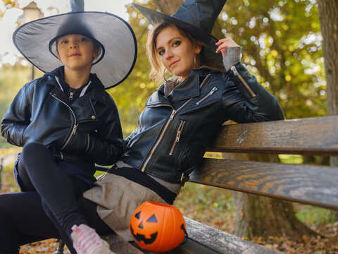 Young mother and daughter sit together on park bench after Halloween preparation, wearing festive costumes and enjoying autumn afternoon surrounded by colorful foliage. in Zagreb