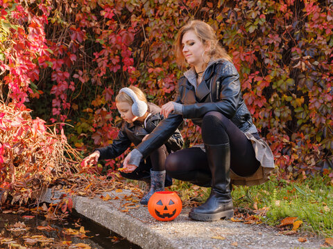 Mother and young daughter pose together in front of vivid red wild grape ivy wall in autumn. Family wears cute Halloween hats on sunny fall day.