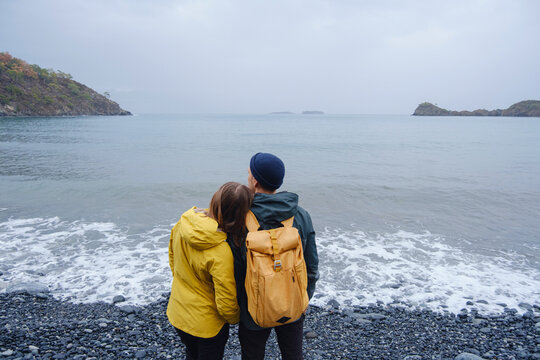 Couple enjoys Turkish coastline. Sitting on stones, watching sea waves, relaxing together, slow romantic moment near calm blue water under soft winter light.