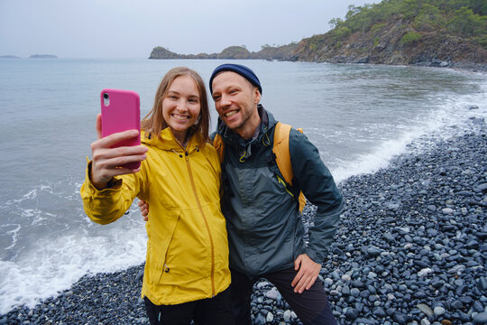 Couple enjoys Turkish coastline and taking selfie on beach in Winter. walking on stones, watching sea waves, relaxing together, slow romantic moment near calm blue water under soft winter light.