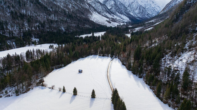 Drone captures majestic snow covered peaks and winding road in Triglav National Park. Traditional wooden cabin sits surrounded by frozen alpine pine forest near Vrsic Pass during winter day.