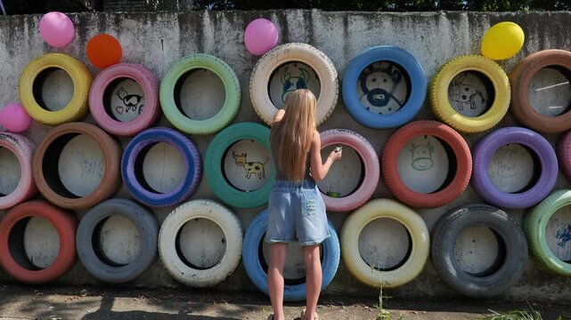 Girl painting colorful cow drawings on recycled tires, creating art on a farm wall