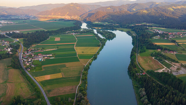 Scenic aerial shot of calm Drava river near Dravograd in Slovenia during blue hour twilight. Soft evening light reflects on water surface surrounded by dark hills and forest.