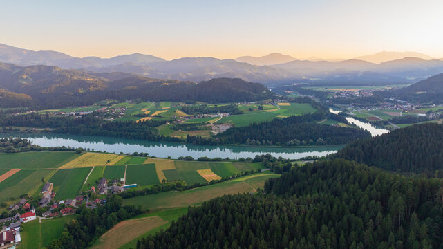 Scenic aerial shot of calm Drava river near Dravograd in Slovenia during blue hour twilight. Soft evening light reflects on water surface surrounded by dark hills and forest.