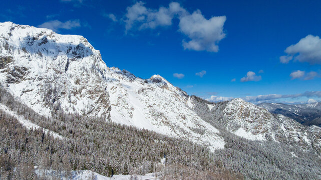 Drone captures majestic snow covered peaks and winding road in Triglav National Park. Traditional wooden cabin sits surrounded by frozen alpine pine forest near Vrsic Pass during winter day.