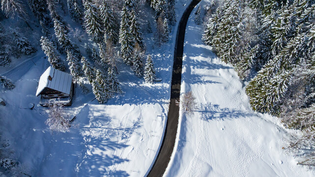 Drone captures majestic snow covered peaks and winding road in Triglav National Park. Traditional wooden cabin sits surrounded by frozen alpine pine forest near Vrsic Pass during winter day.