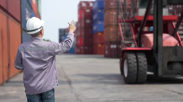 Rear view of a logistics officer walking between two shipping containers toward a reach stacker while checking his paperwork during a routine shift at the container terminal.