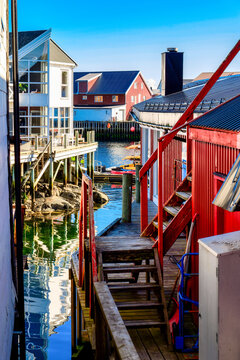 Narrow harbor alley in Henningsv&aelig;r on the Lofoten Islands between traditional red and white wooden houses, wooden walkways and stilt buildings with calm water and reflections.