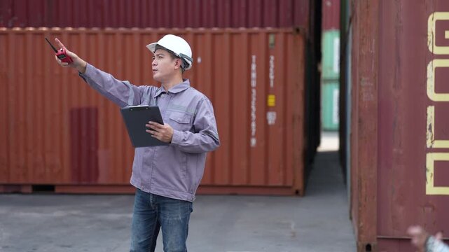 A yard foreman speaks into a handheld radio and looks up at the cargo boxes while holding an inspection clipboard at his side at a busy container terminal yard.