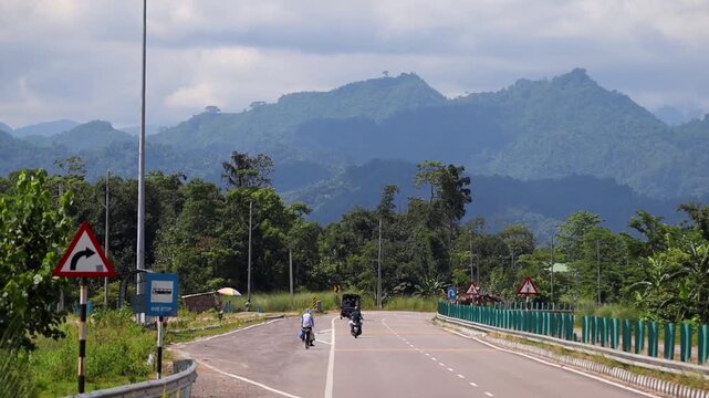 Dibrugarh, Assam, India &ndash; 23 September 2026: A scenic roadway winding through lush green vegetation with motorcycles and a three-wheeler traveling along the road