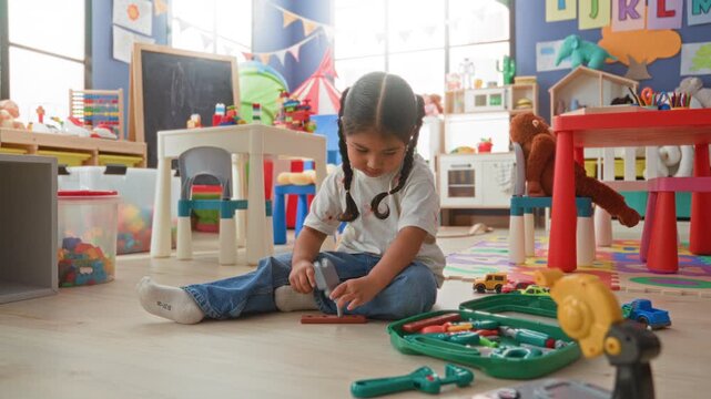 Girl sits on floor playing with a green toy toolkit and small cars on a foam playmat in a preschool classroom; quiet concentration learning.