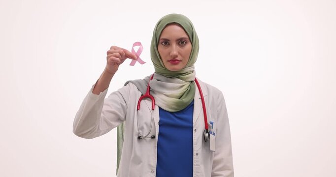 Doctor in hijab holds delicate pink ribbon symbol of breast cancer in hands. Woman promotes important message about health of women at white wall