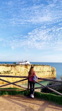 woman leaning on fence looking at nossa senhora da rocha chapel on cliff algarve portugal
