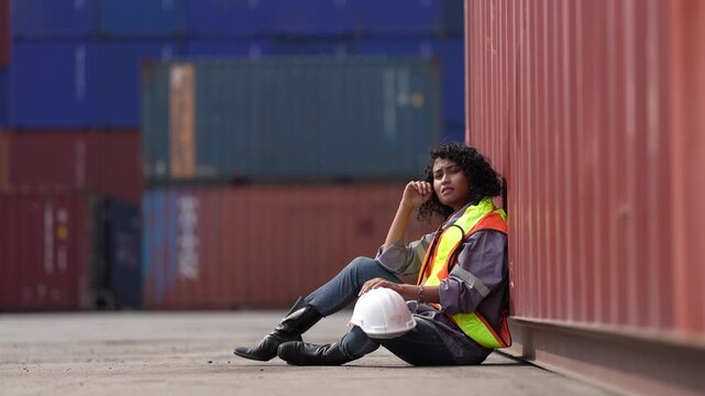 A tired female dock worker sits on the ground leaning against a shipping container, resting her head on her hand during a break at a busy container terminal yard.