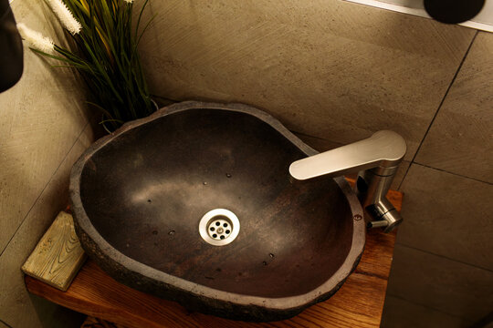 dark stone oval washbasin with chrome faucet on wooden countertop, tiled wall and green plant behind, modern bathroom interior, no people, design concept, minimalist restroom in natural light