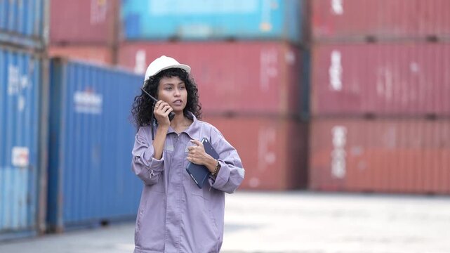 Female logistics worker wearing a safety hard hat holding a digital tablet while talking on a walkie talkie radio and walking outdoors at a freight shipping container yard terminal facility