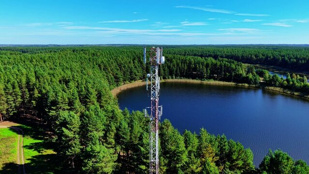 Cell tower surrounded by lush green forest and calm blue lake under clear skies