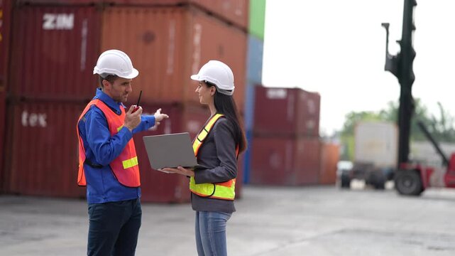 A supervisor calls the crane operator on his radio while a female colleague holds an open laptop with the container loading list at a busy container terminal yard.