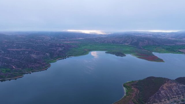 Vail Lake spreads between rugged hills beneath a low cloud layer and muted overcast sky, aerial panoramic ascend into misty clouds