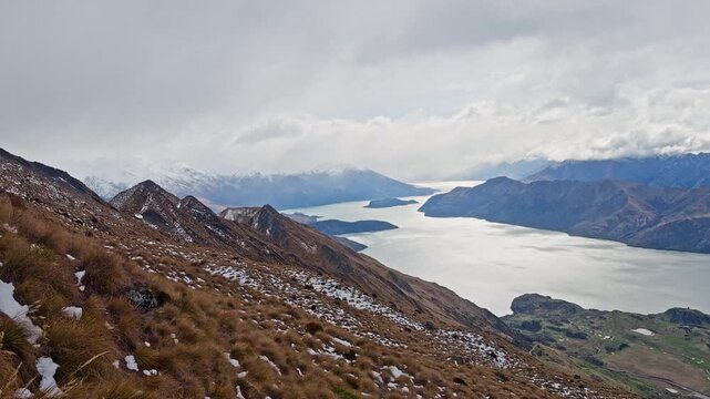 Alpine hiking trail with snow patches overlooking Lake Wanaka and distant mountains